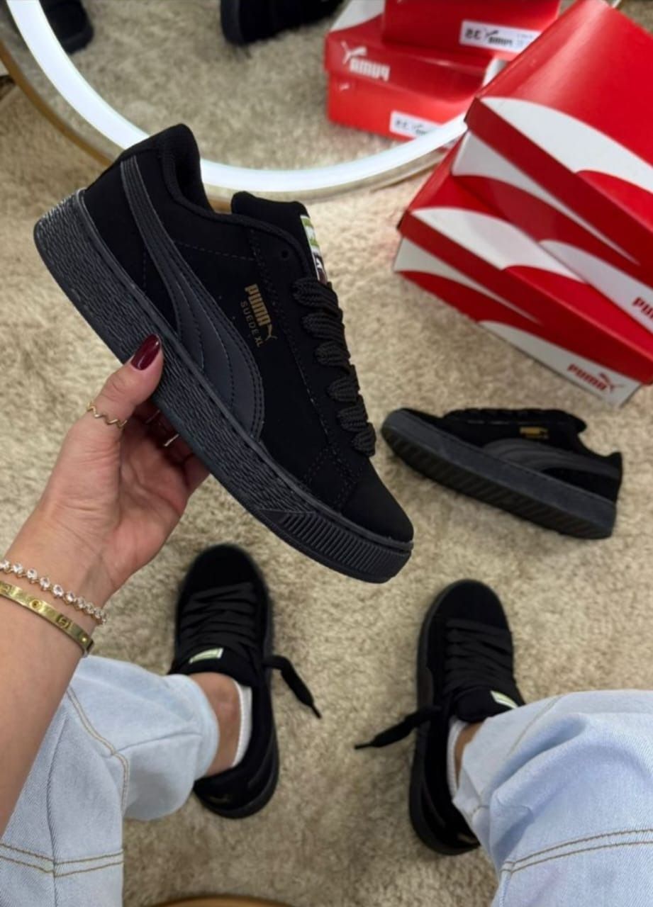 Persona sosteniendo una zapatilla deportiva Puma Suede negra, otros pares visibles, junto a una caja de zapatos.