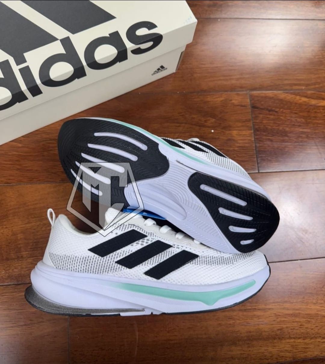 Zapatillas Adidas blancas y negras con detalles en menta, cerca de una caja de Adidas sobre una superficie de madera.
