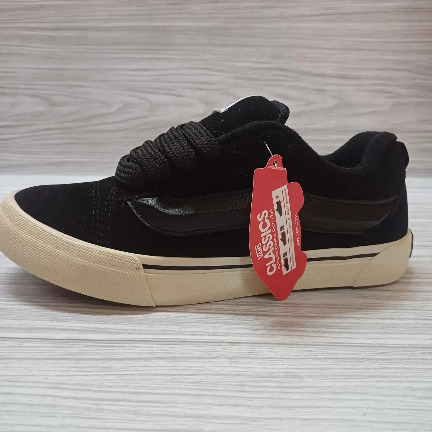 Zapatilla Vans negra con suela color crema y etiqueta roja, sobre una superficie de madera clara.