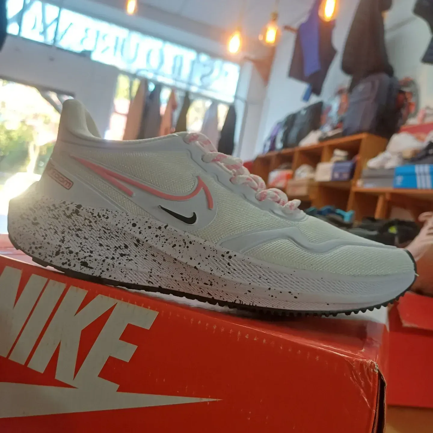 Zapatilla Nike blanca para correr con detalles rosas, sobre una caja de zapatos naranja en una tienda.