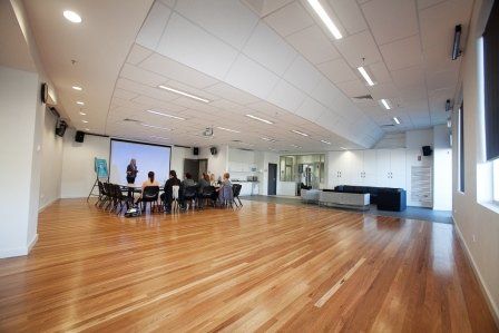 Large spacious social hall with wooden floors . Group having a meeting  using tables and chairs with a presentation on projector screen.