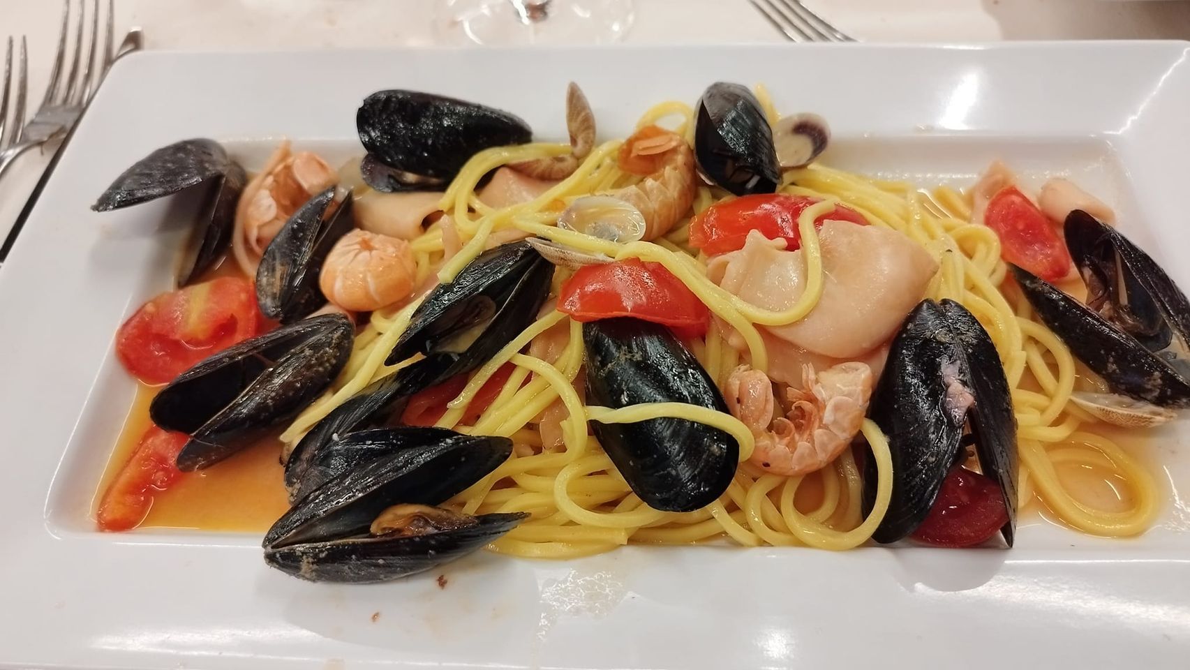 Spaghetti con cozze, gamberi, calamari e pomodori su un piatto bianco rettangolare.