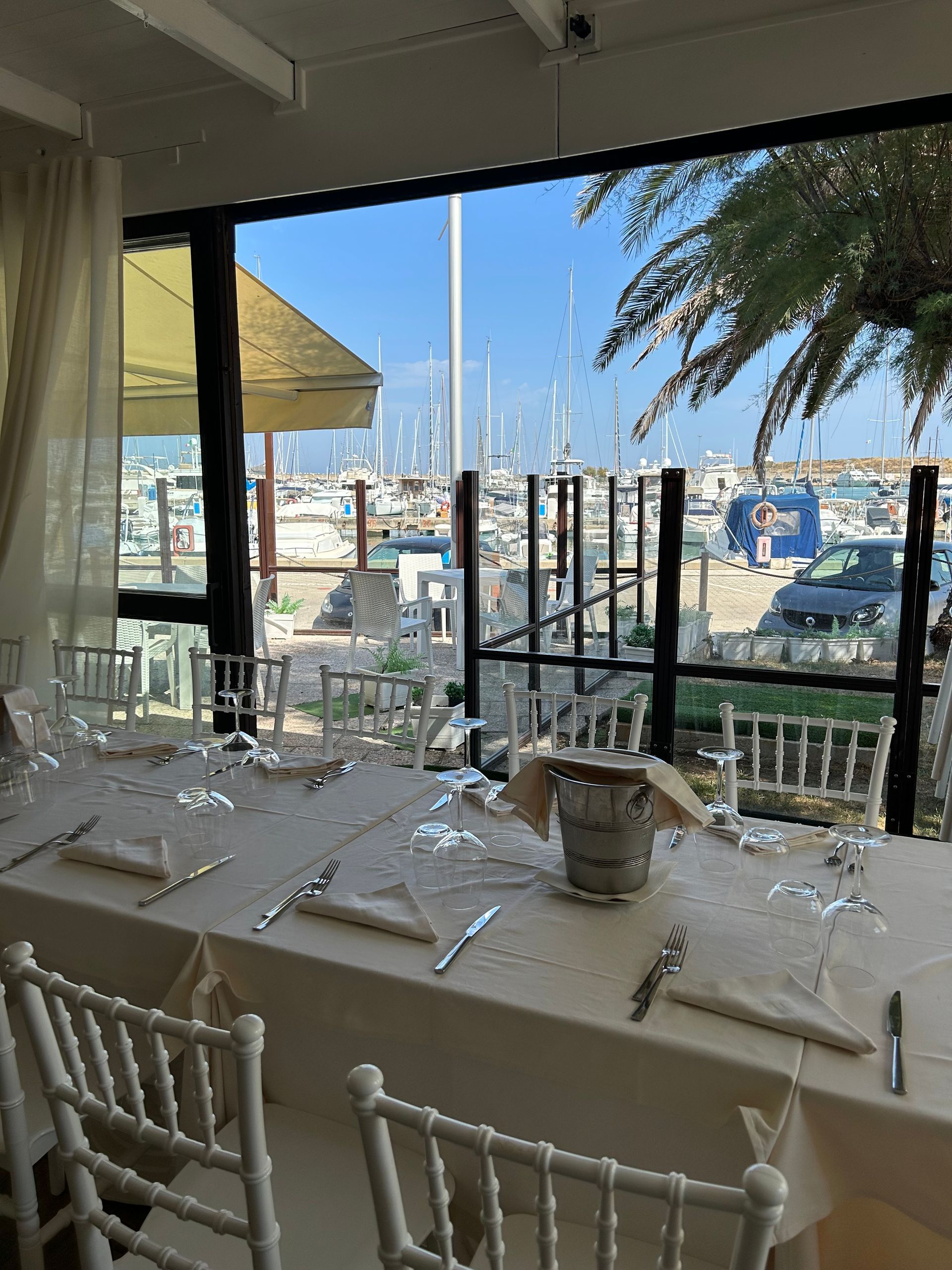 Tavolo di un ristorante apparecchiato per un pasto, con vista sul porto con le barche in una giornata di sole.