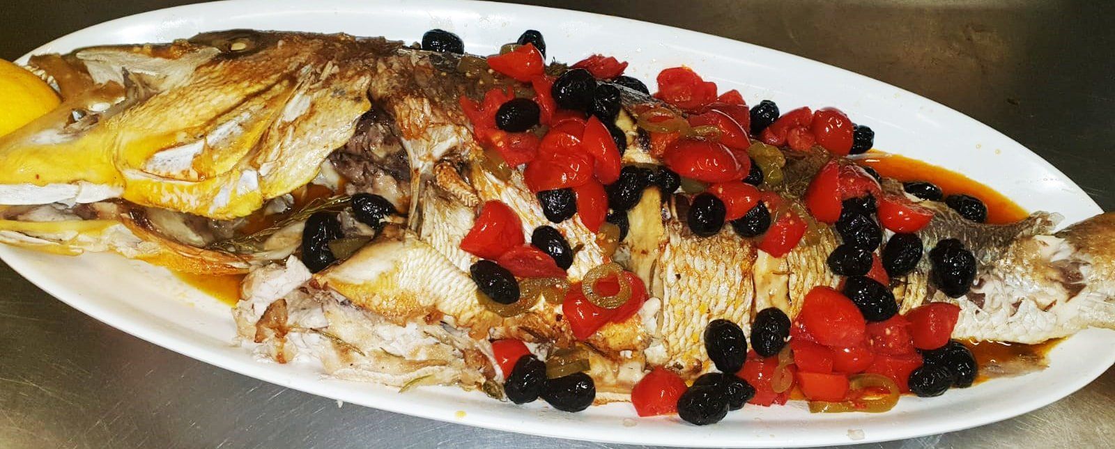 Pesce intero al forno condito con pomodori e olive su un piatto bianco.