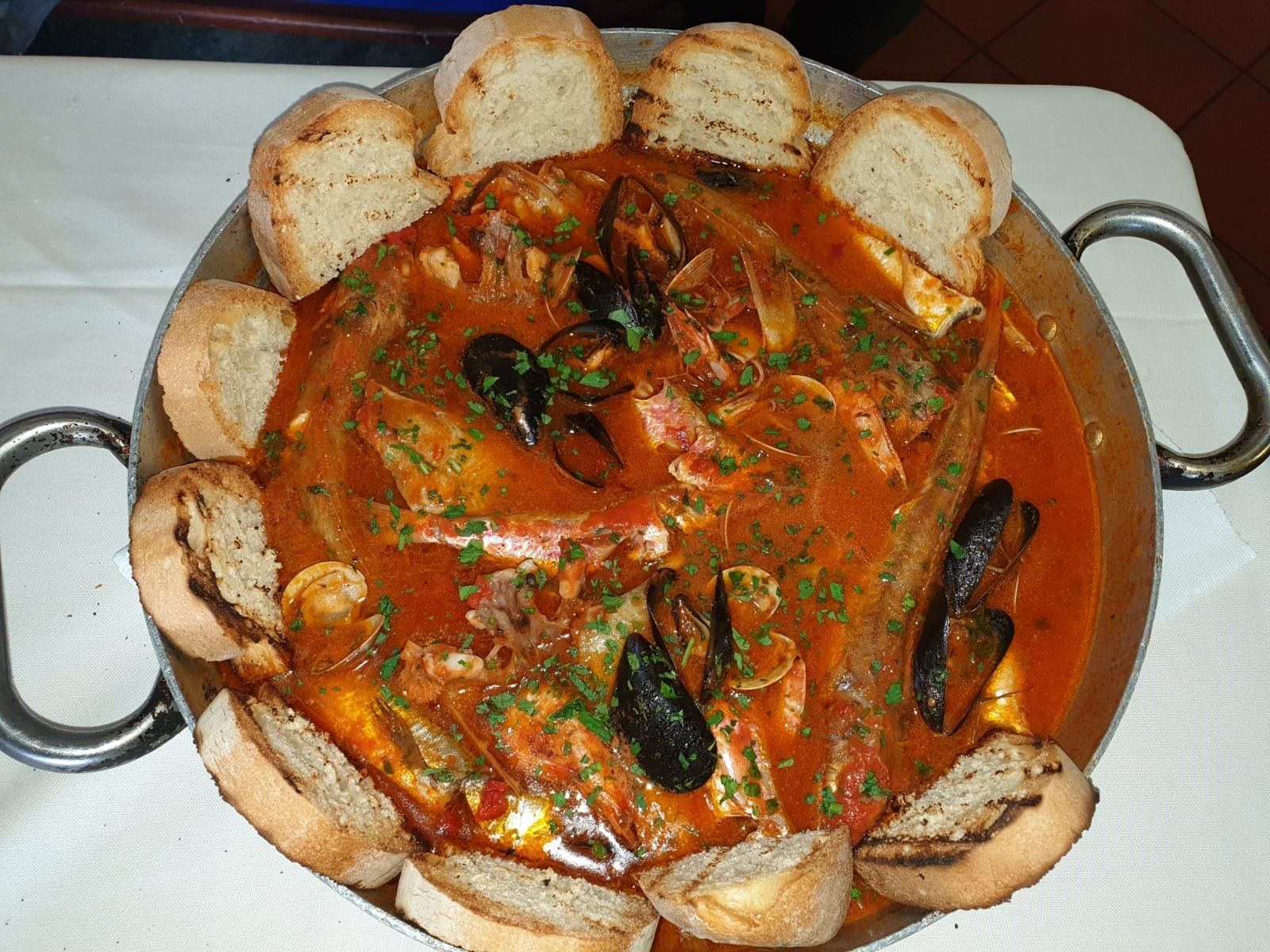 Stufato di pesce in una padella di metallo, circondato da fette di pane tostato.