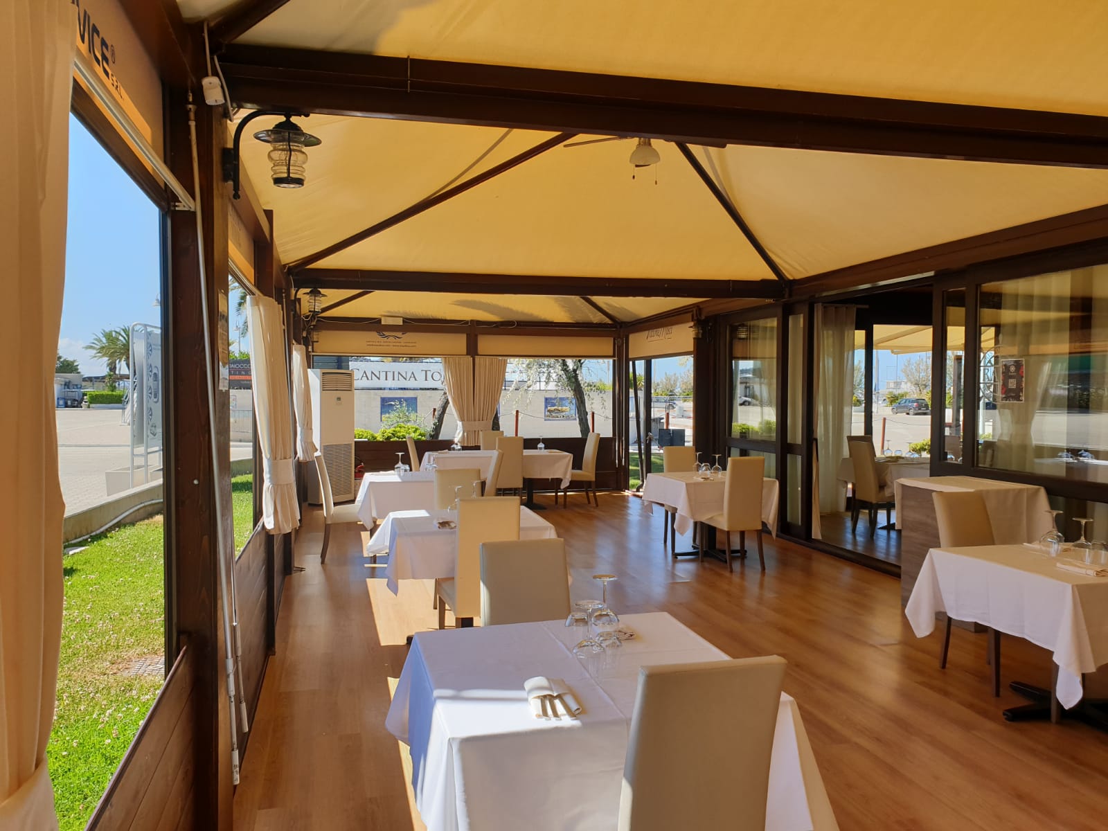 Ristorante con tovaglie bianche, tende color cuoio, travi in legno, vetrate e vista soleggiata.