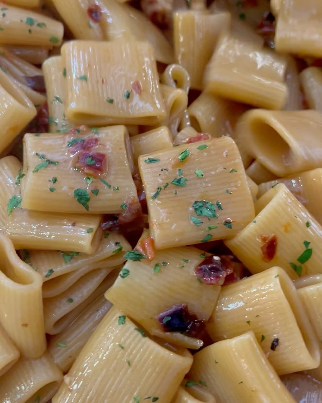 Primo piano di pasta rigatoni condita con una salsa leggera, con pezzetti di erbe aromatiche e ingredienti rossi.