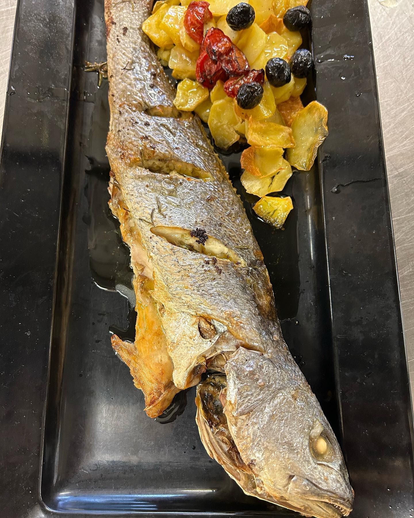 Pesce intero fritto con patate arrosto, olive e pomodori su un piatto nero.