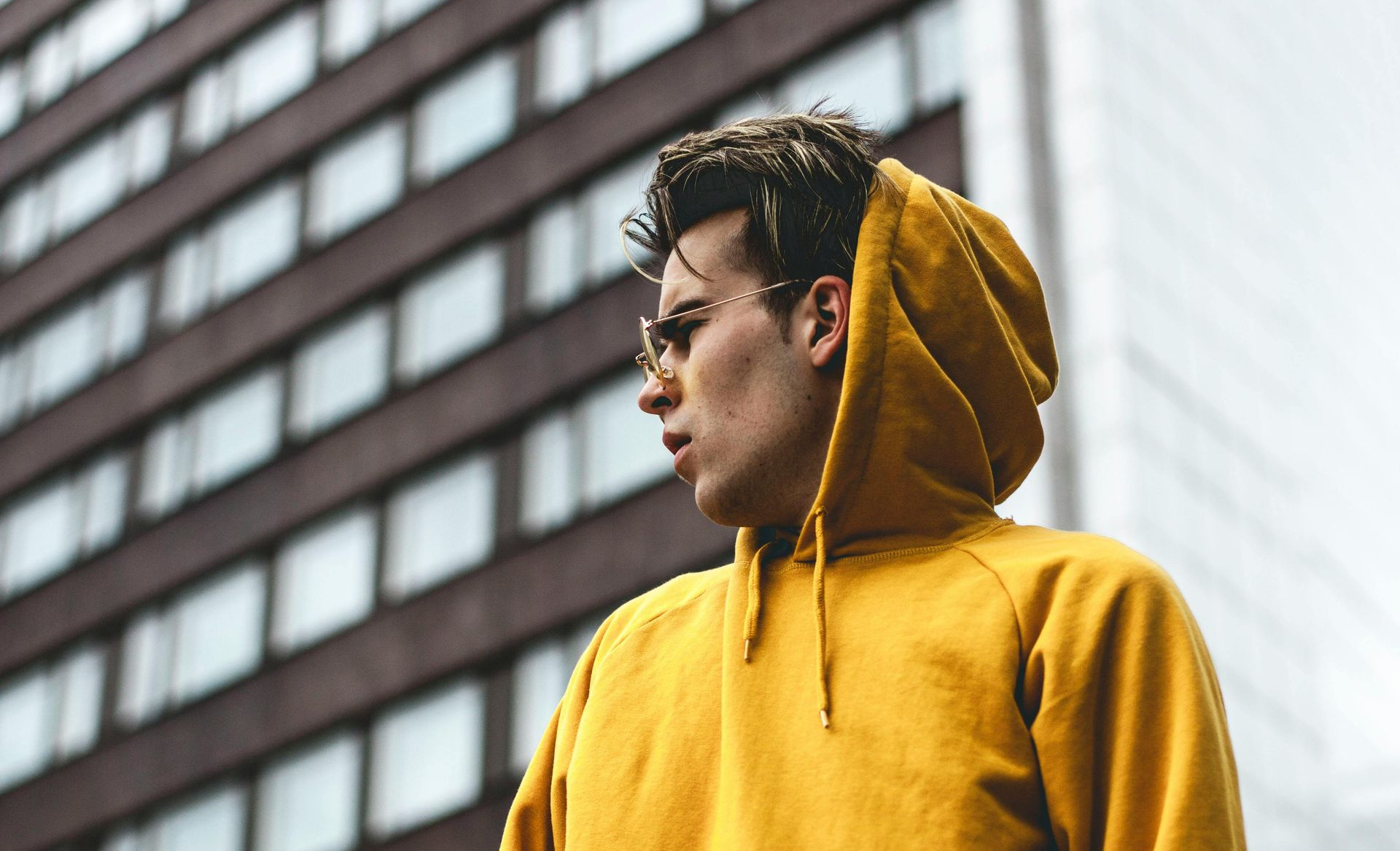 Un uomo con una felpa gialla e occhiali da sole distoglie lo sguardo dalla telecamera. Sullo sfondo un edificio marrone.