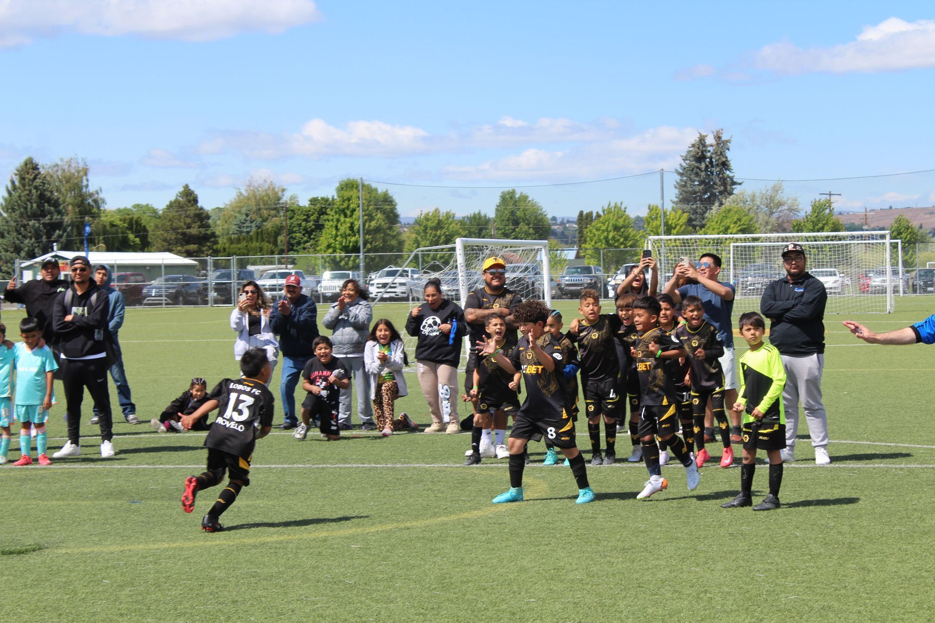 A youth soccer team in black uniforms celebrates on a field while parents and coaches watch from the sidelines.