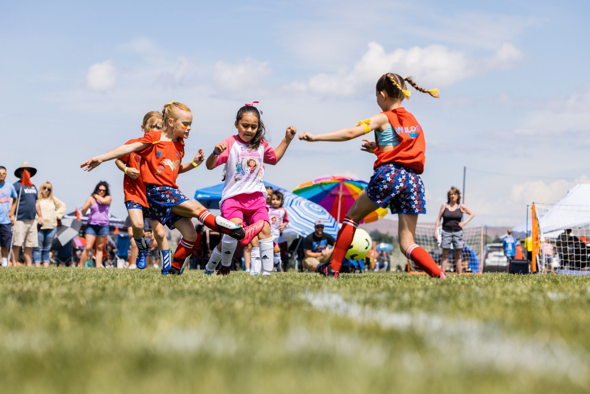 Children in colorful jerseys playing a soccer match on a sunny field, with spectators in the background.