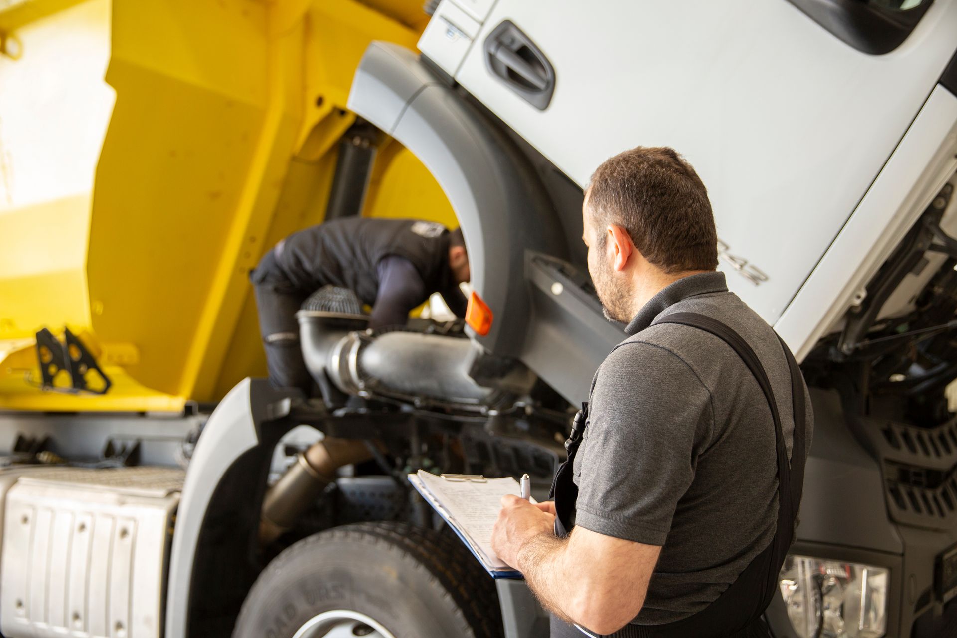 Mechanic inspects truck engine with hood open while another works under the cab.