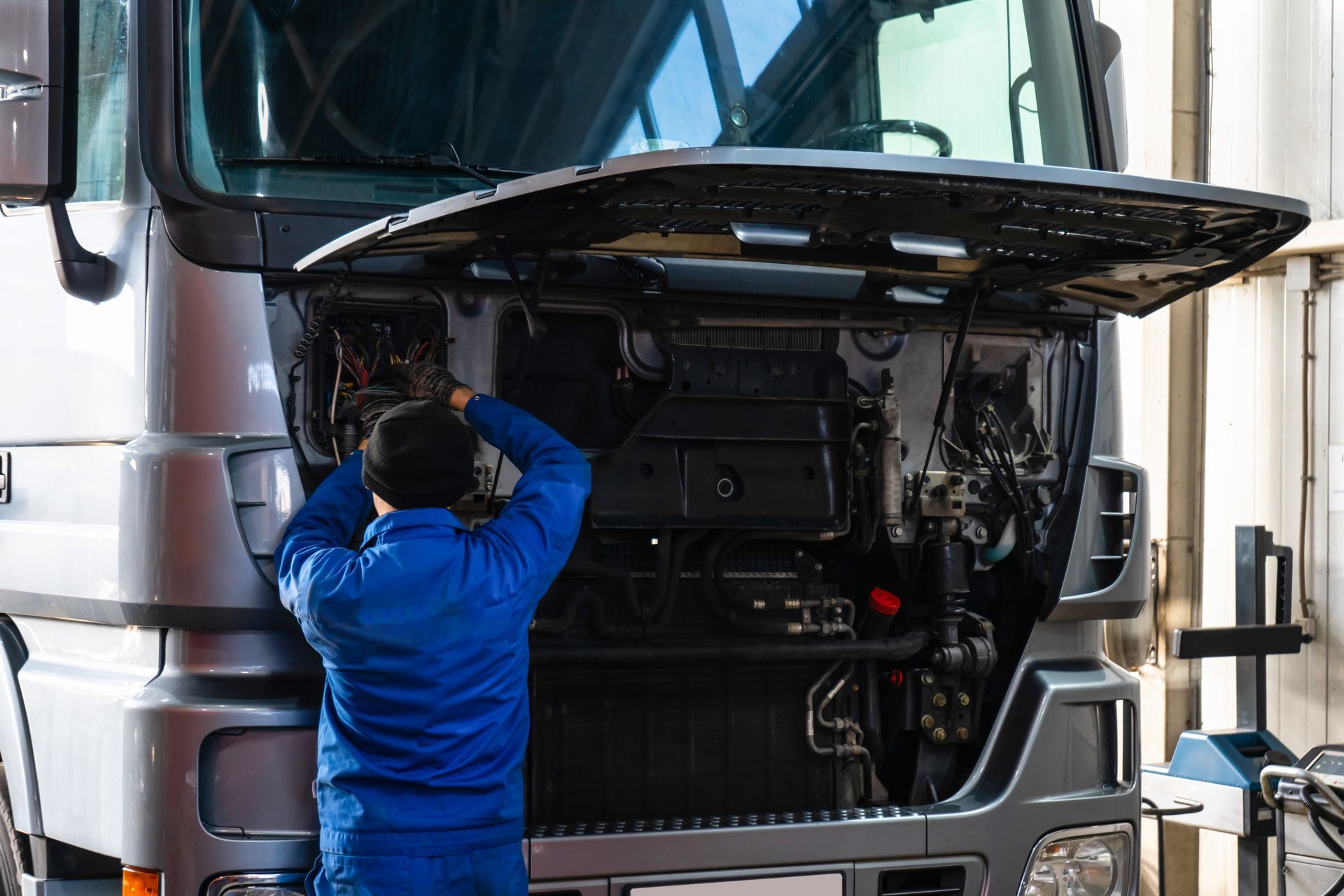 Mechanic in blue uniform working on wiring under the open hood of a large truck.