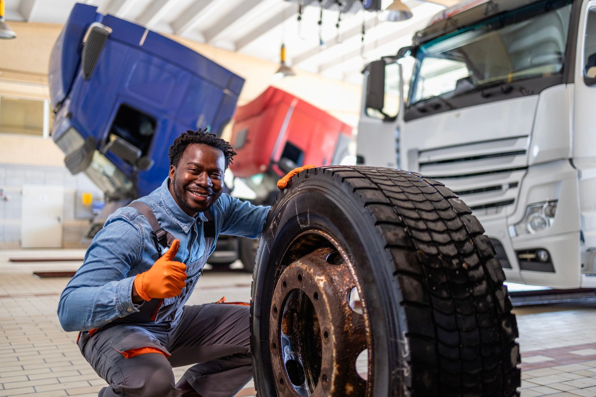 Mechanic kneeling by a large truck tire in a repair shop with trucks in background.