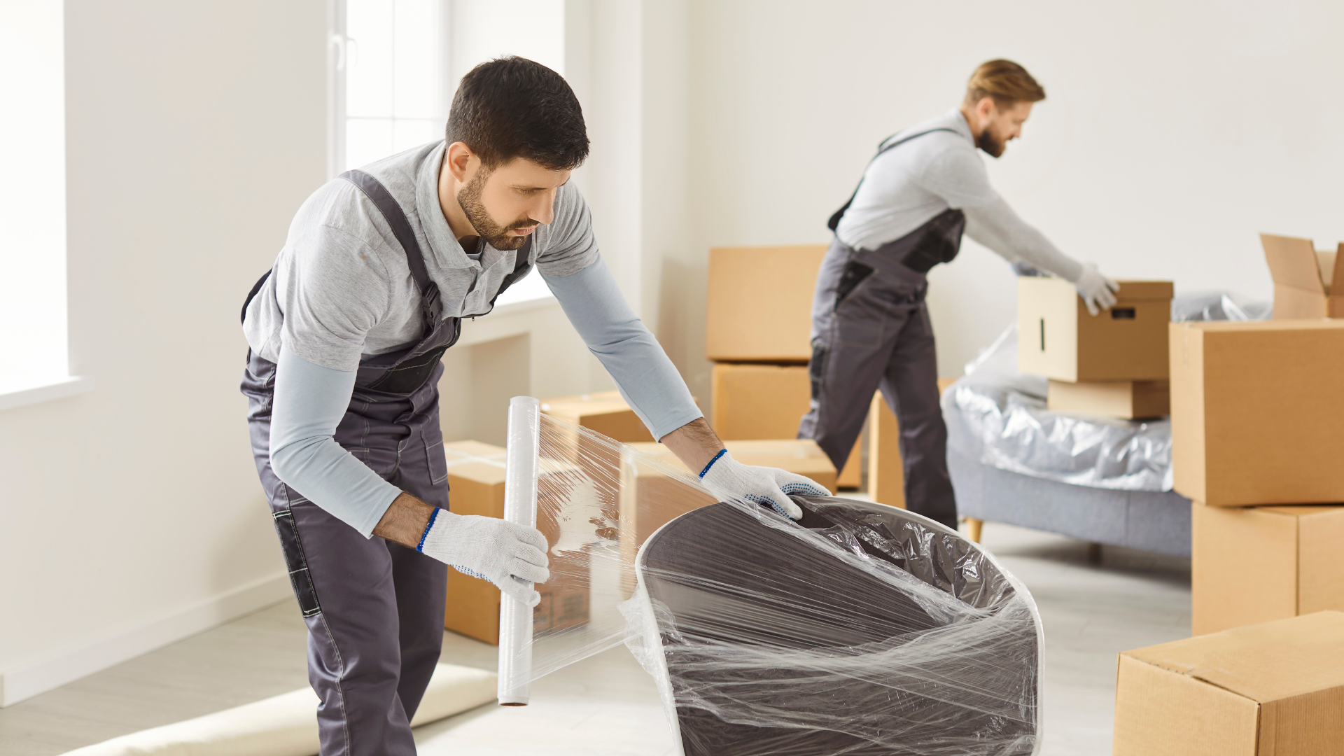 Two professionals in grey uniforms pack items with bubble wrap and cardboard boxes in an unfurnished room.