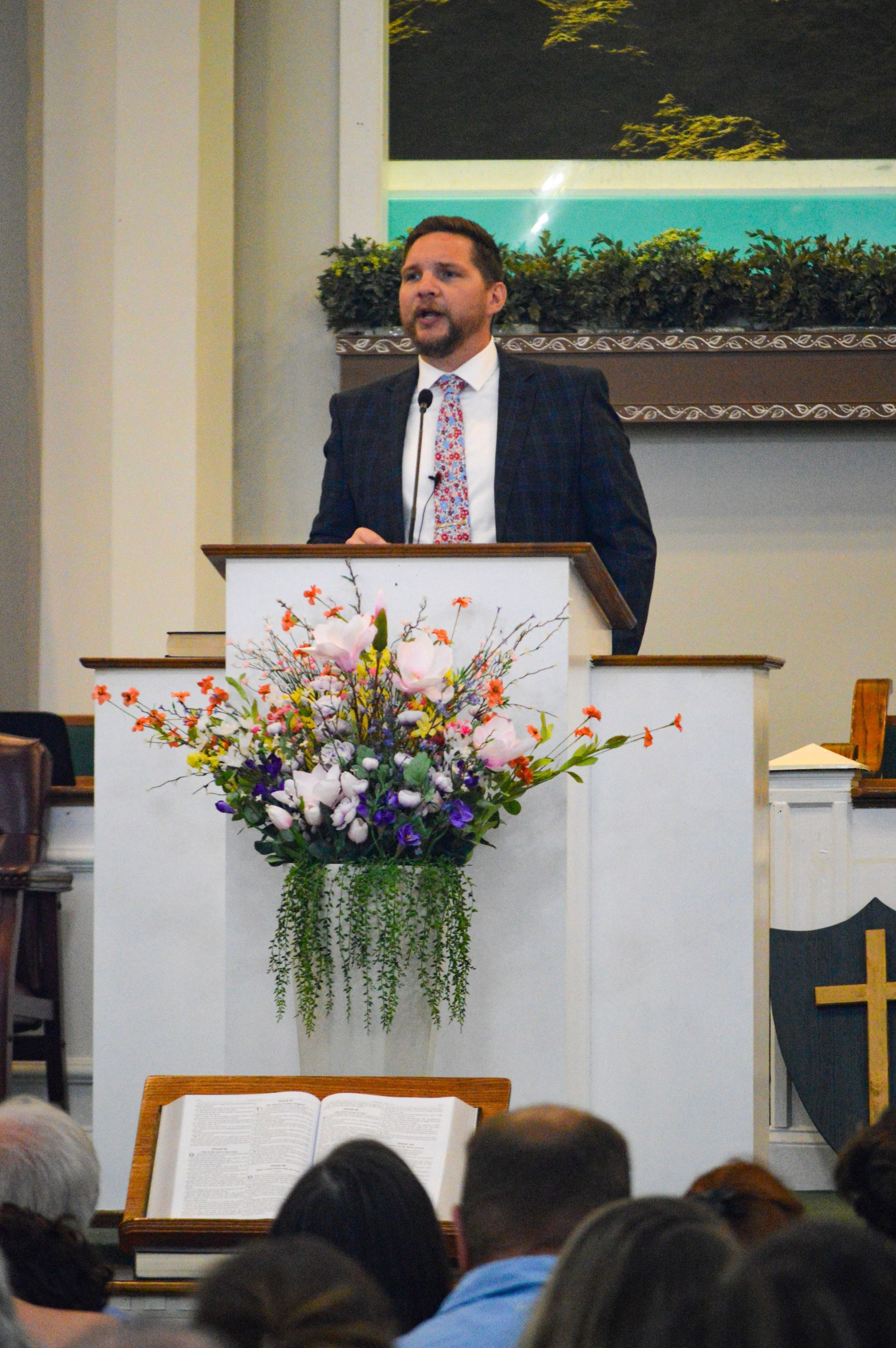 Man in a suit, glasses, and tie smiles at the camera, set against a gray background.