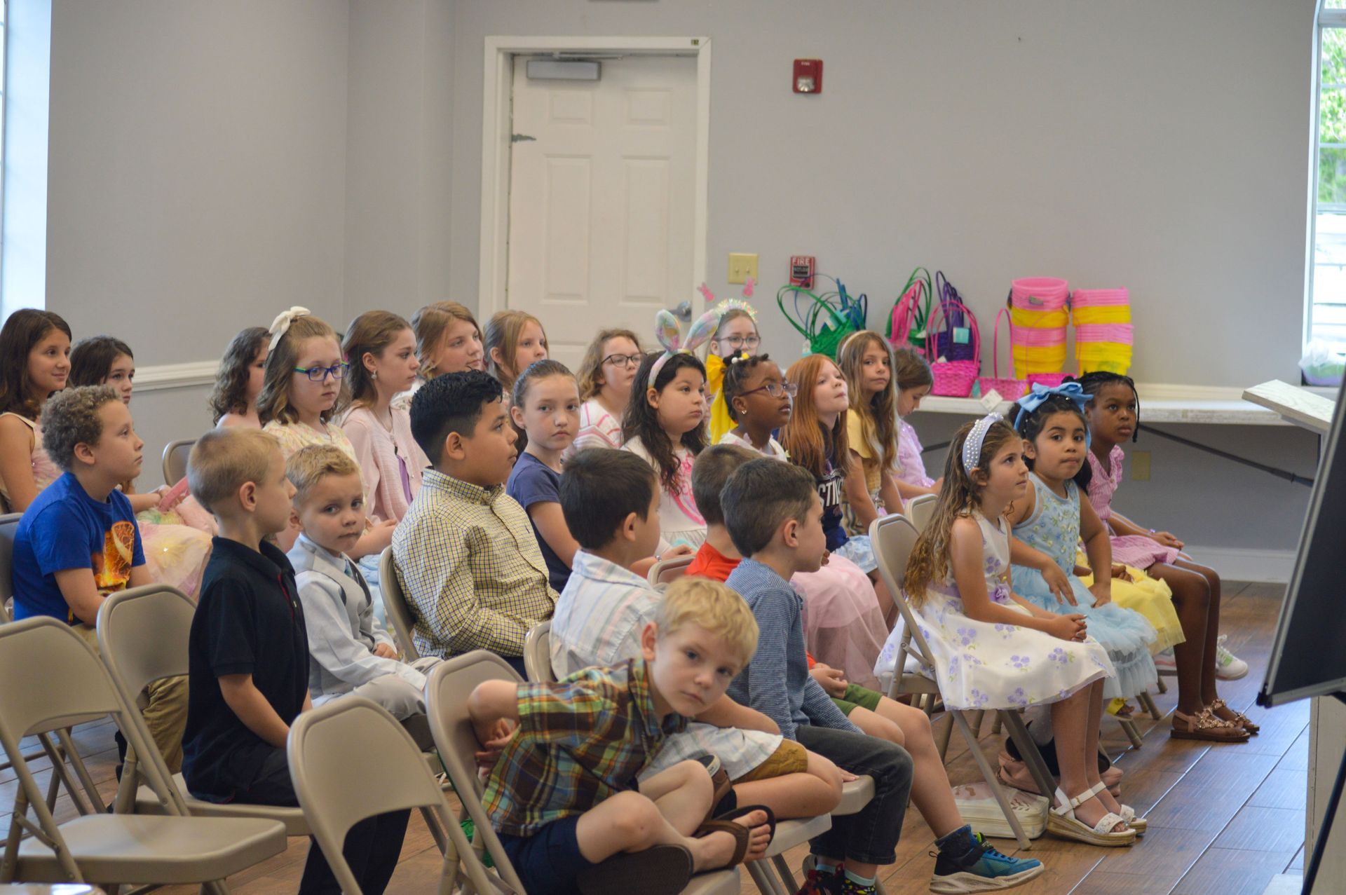 A man lecturing a group of children in a classroom; children sitting, listening; white and light wood room.