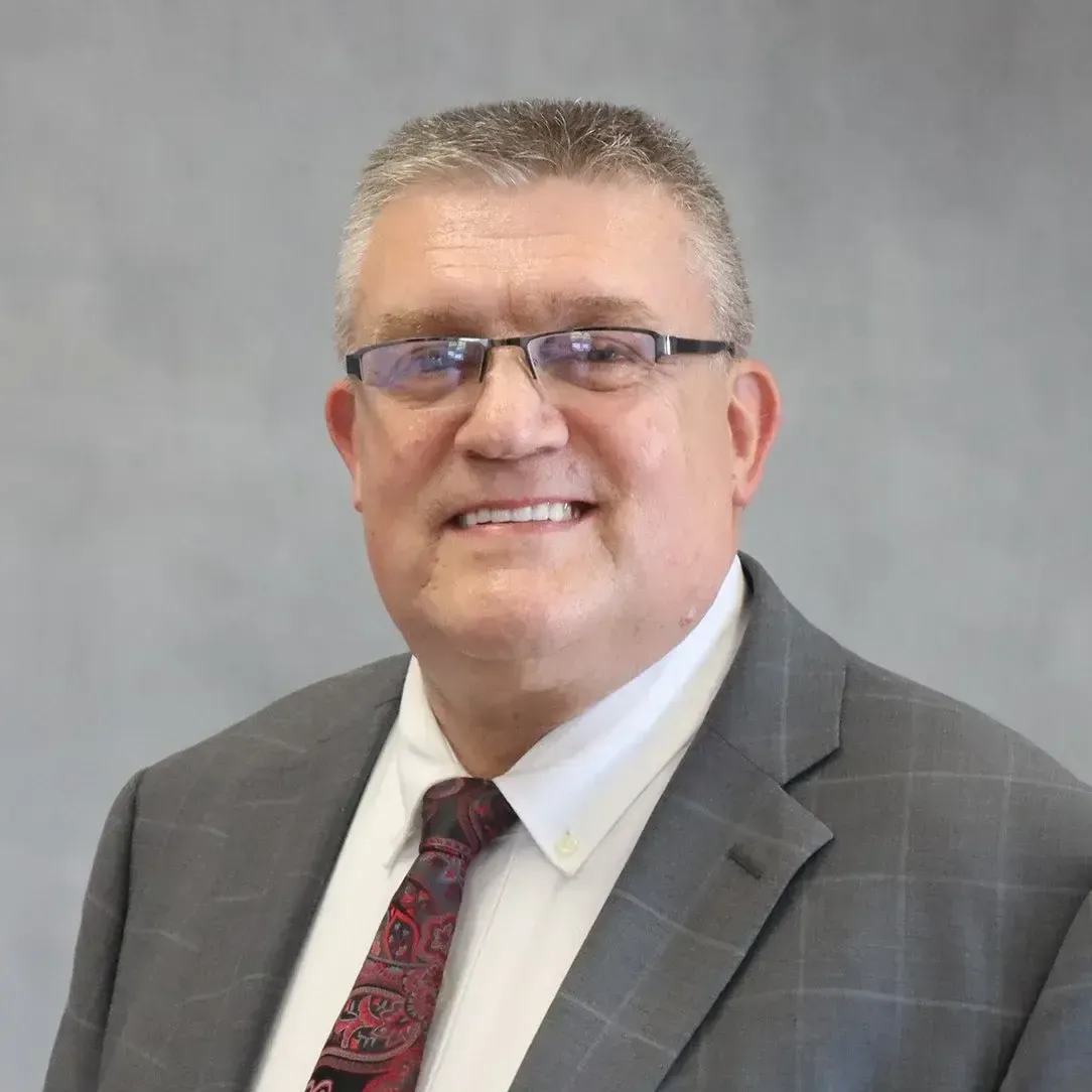 Man in glasses and suit smiles at camera. Gray background.