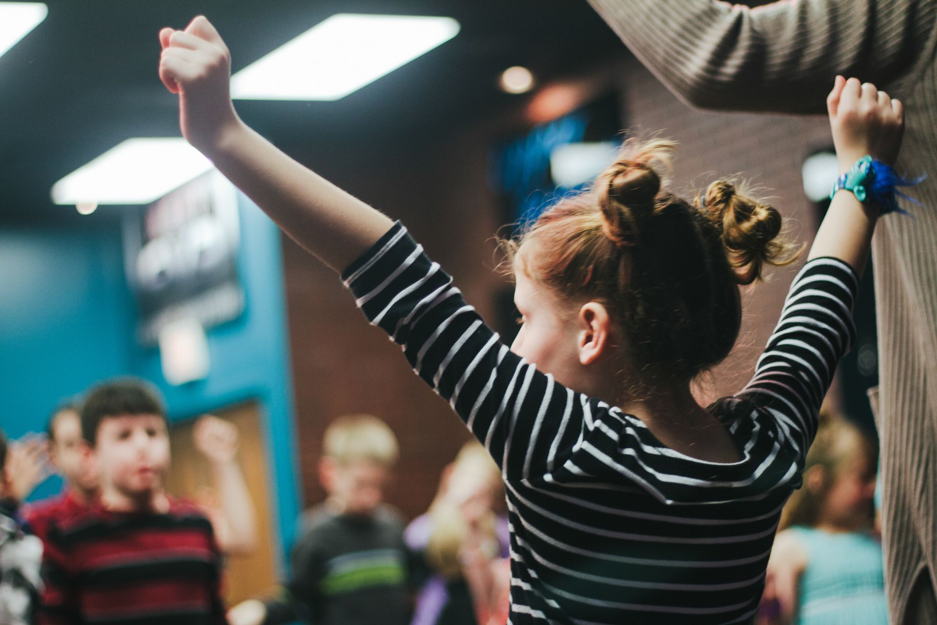 Girl with red hair and striped shirt raises arms in a room full of children.