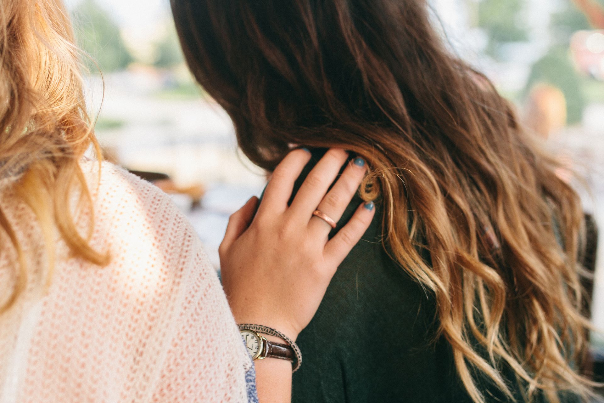 Woman comforts another, hand on shoulder; tan sweater, dark green top, wavy brown hair.