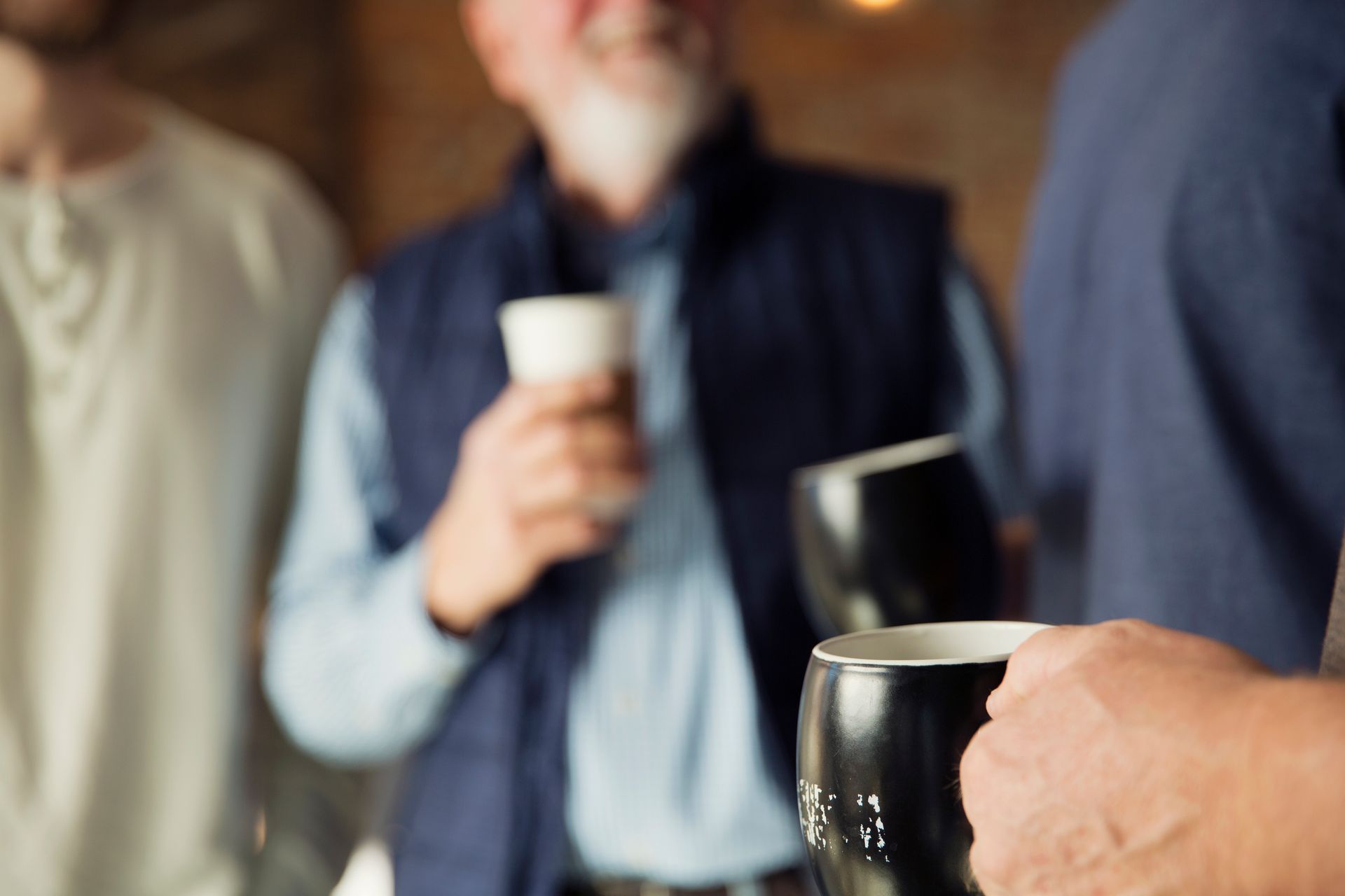 People holding mugs, one man with a drink, blurred background, indoor gathering.