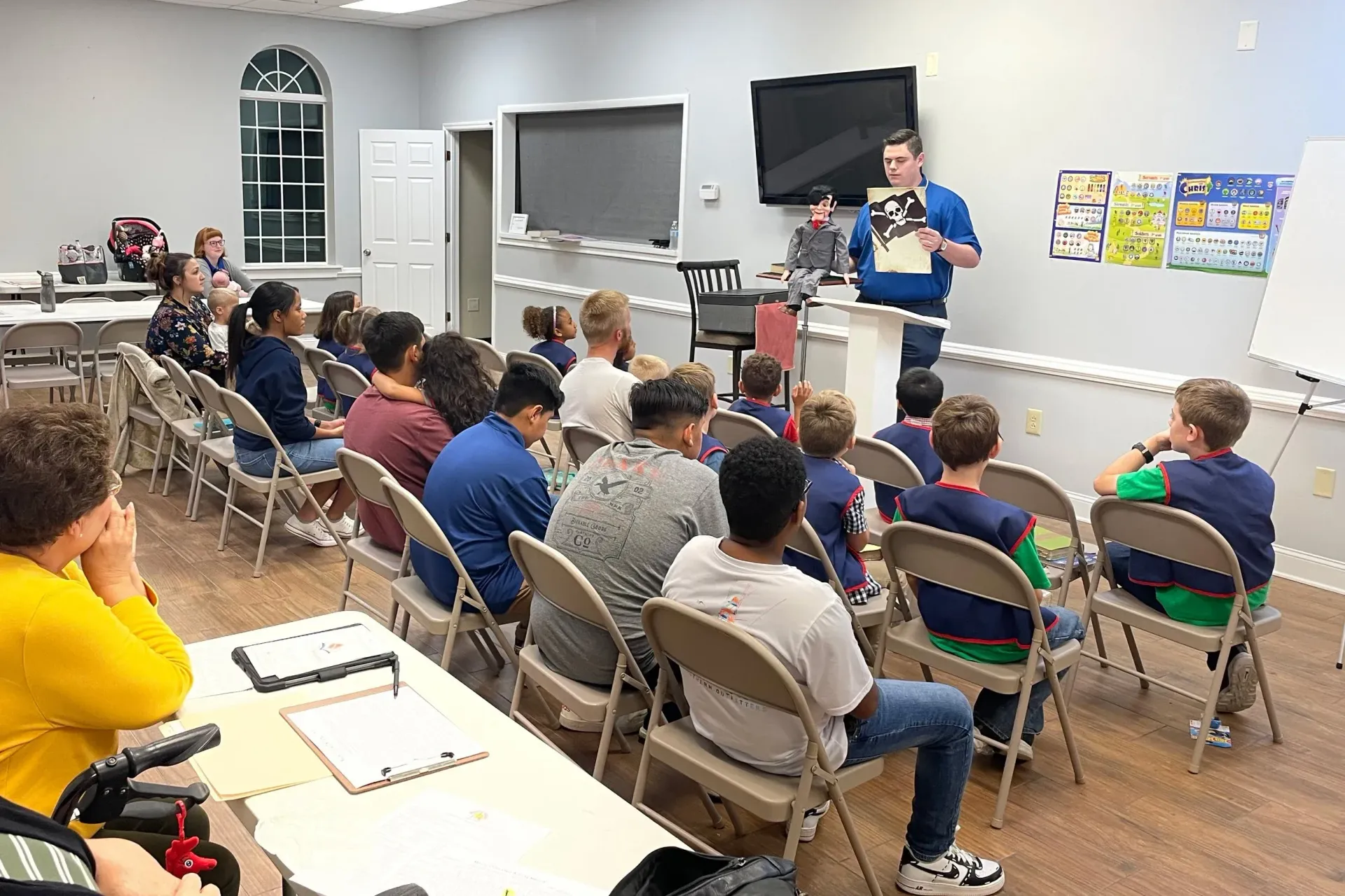 A man lecturing a group of children in a classroom; children sitting, listening; white and light wood room.