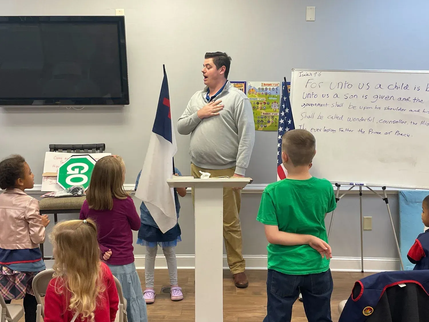 Man leading children in pledge, holding a Christian flag in a classroom setting.