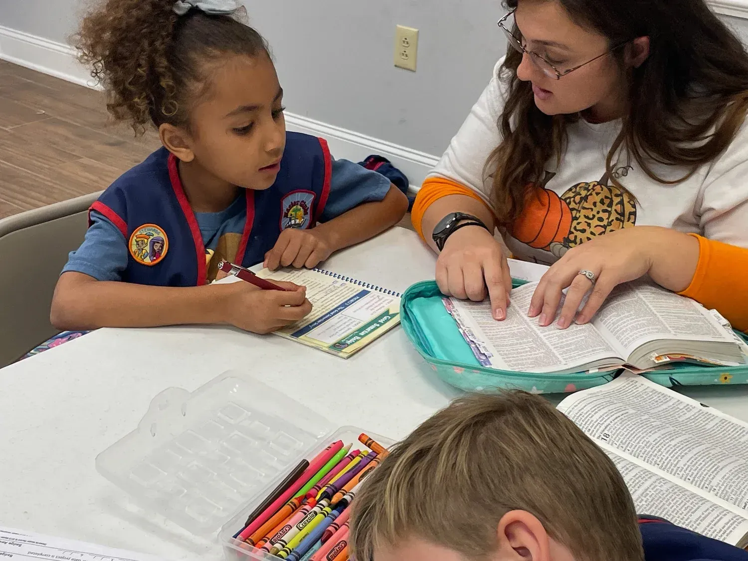 Woman points at Bible, helping girl write in notebook, with boy drawing nearby.