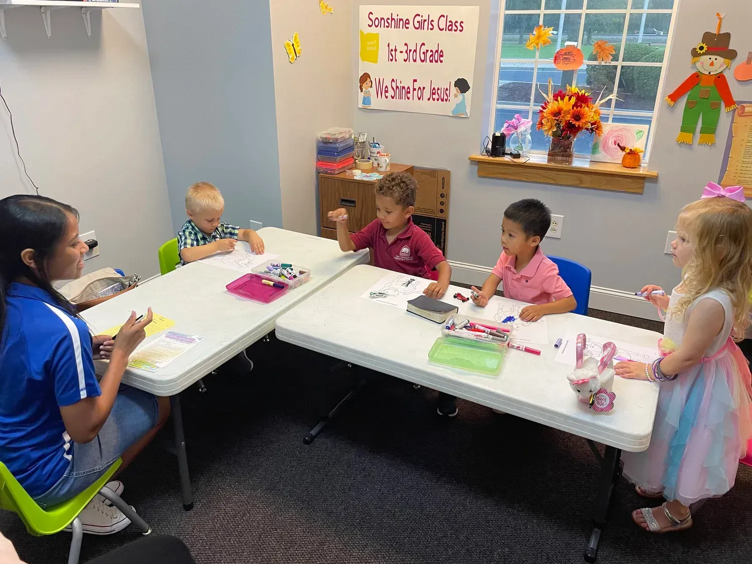 A teacher guides four children at a table, drawing and painting in a classroom setting.