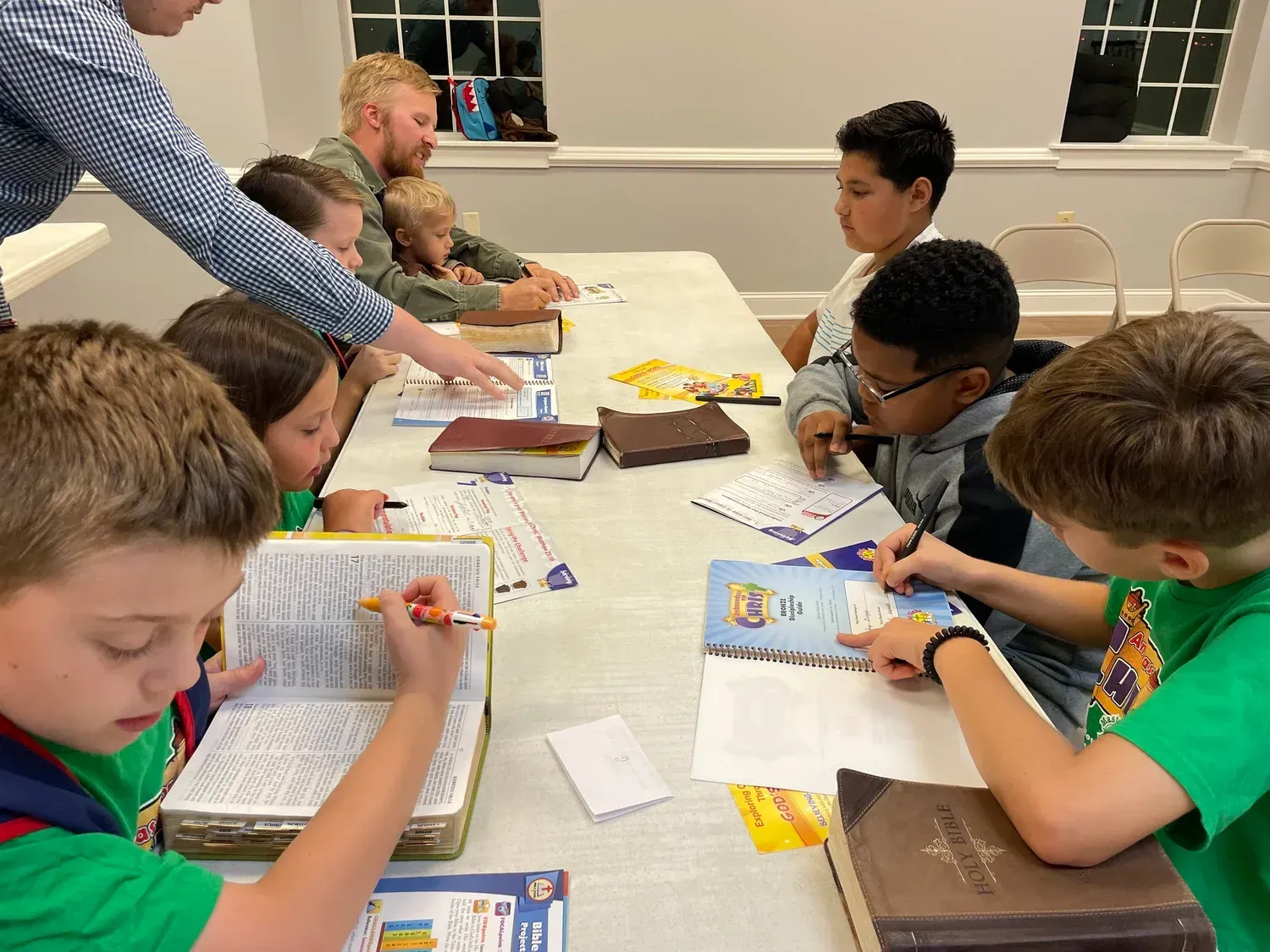 Children and adults at a table, studying books. A white room with warm lighting.