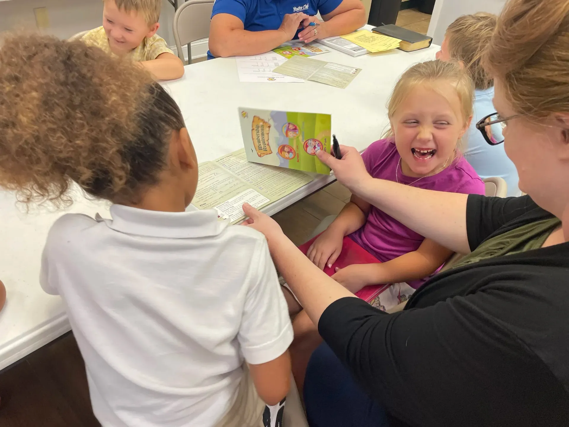Teacher reading a book to two laughing children at a table. Children are seated.