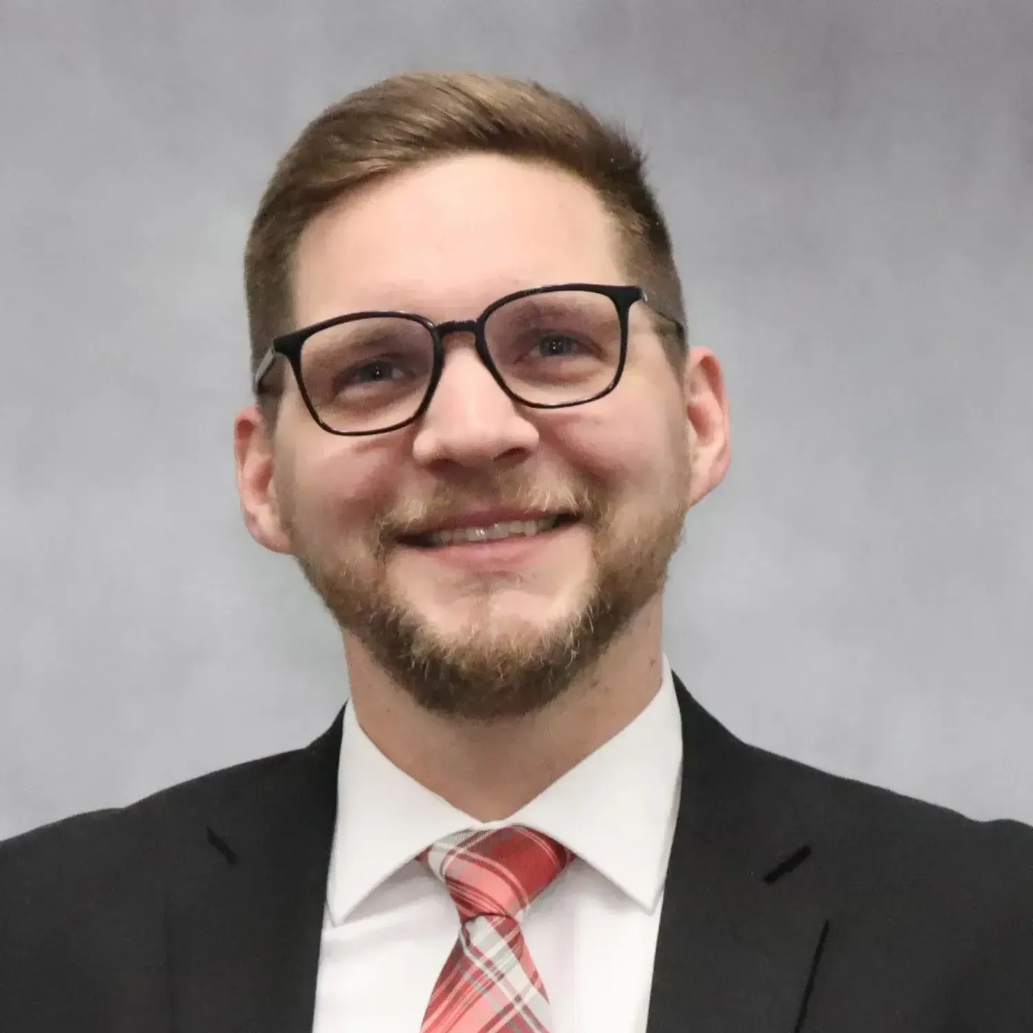 Man in a suit, glasses, and tie smiles at the camera, set against a gray background.