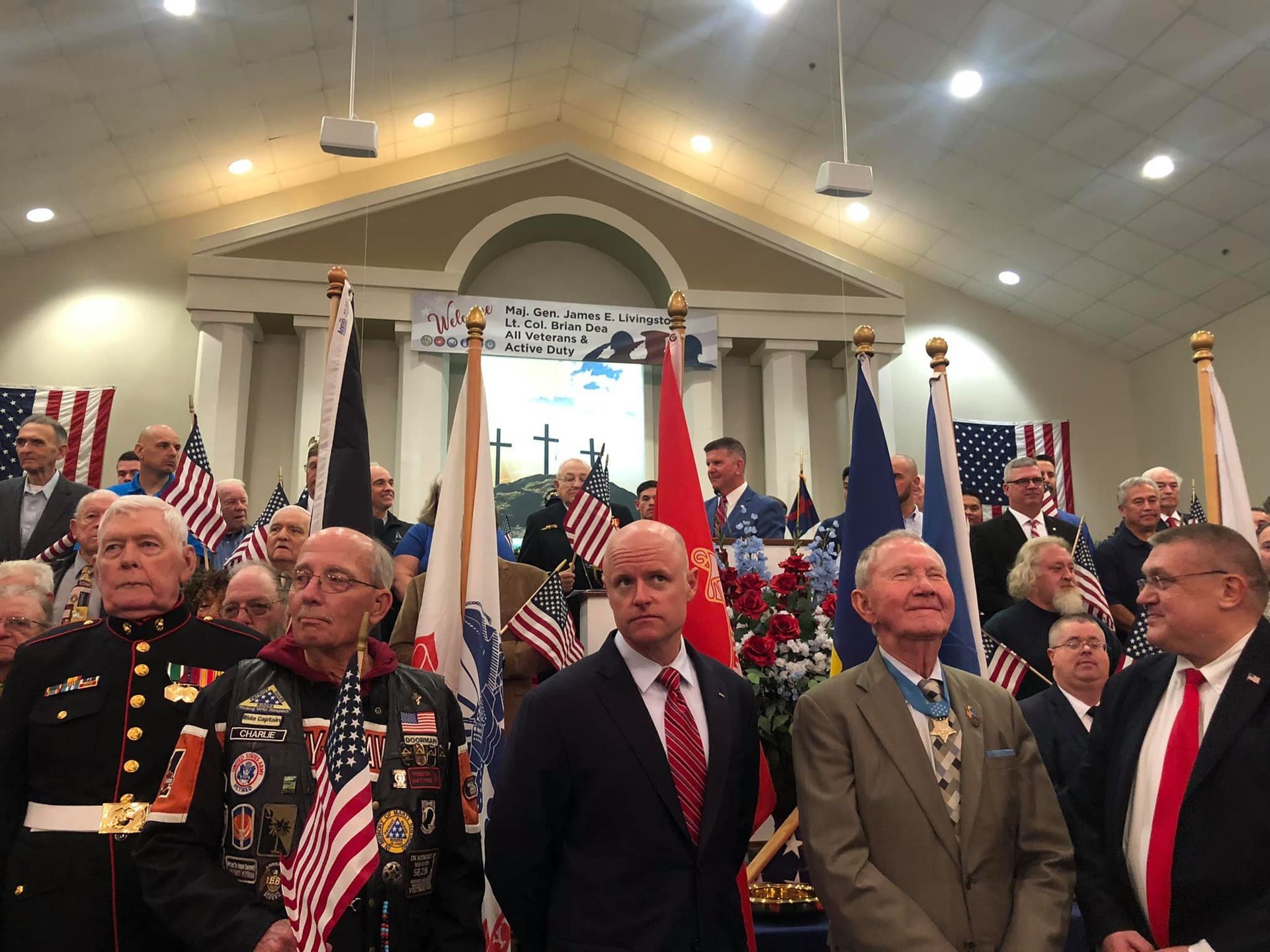 Veterans in a church, some holding flags, listening to a speaker. Red, white, and blue decorations.