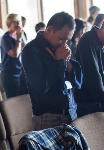 Man with hands clasped in prayer, head bowed, in a room with others.
