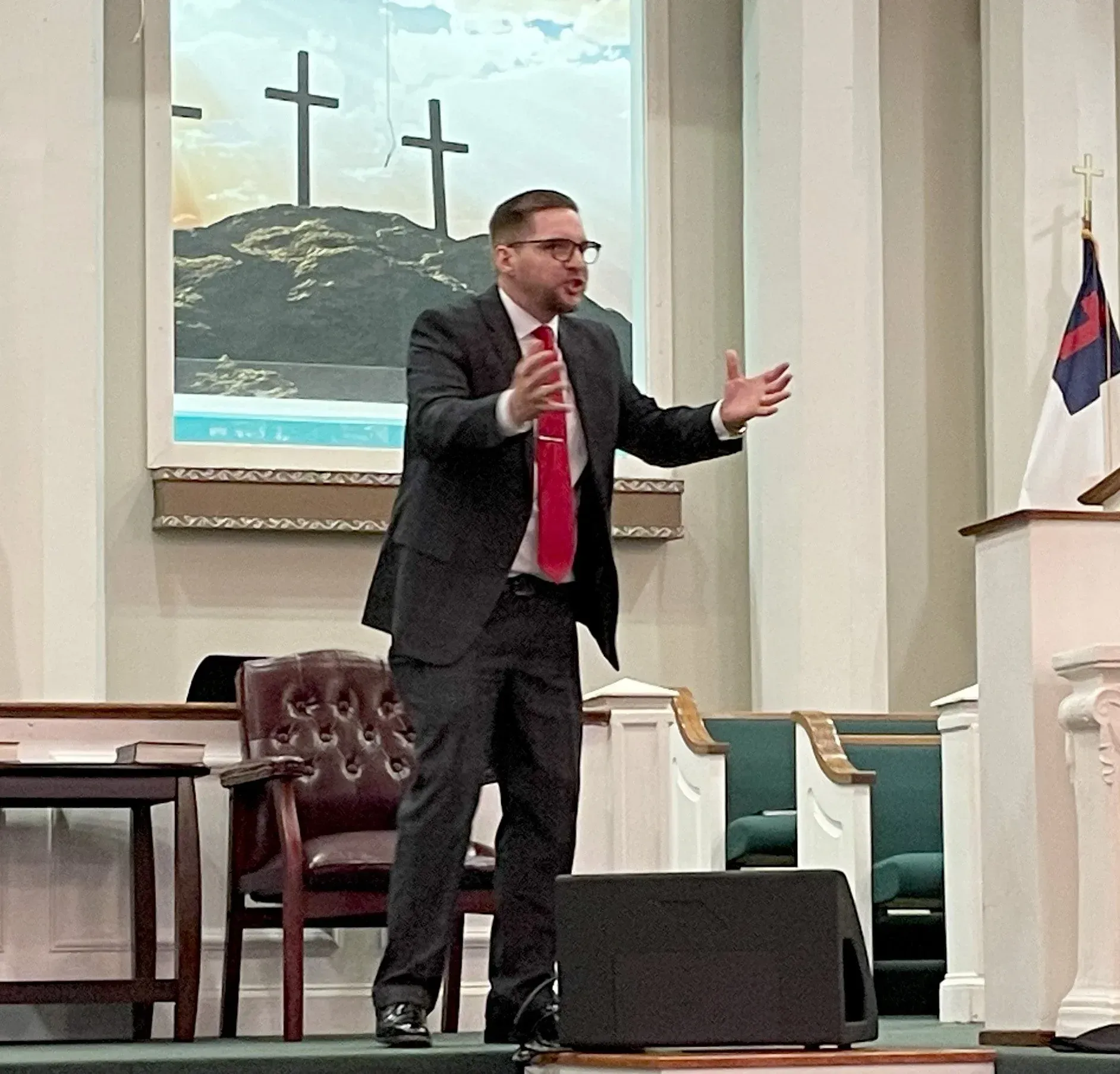 Man in suit, red tie, speaking on stage, gesturing, in church. Cross painting backdrop.