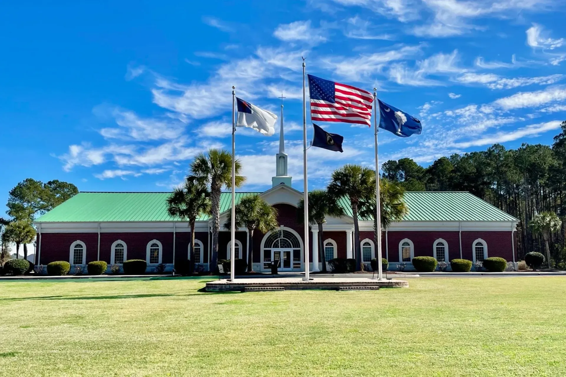 Trident baptist Church building with flags: Cross, US, military, SC. Red brick, green roof, blue sky, grassy lawn.