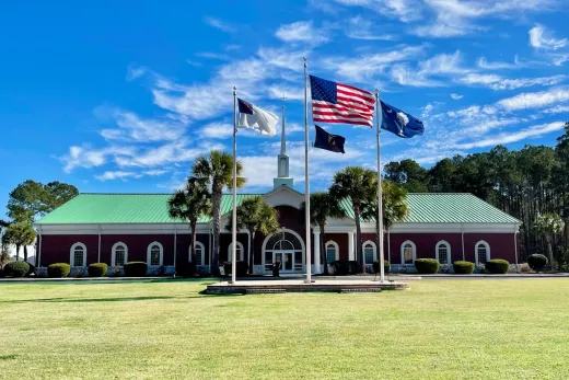 Trident baptist Church building with flags: Cross, US, military, SC. Red brick, green roof, blue sky, grassy lawn.