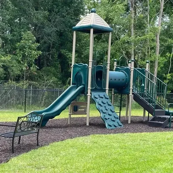 Playground with green slide, climbing wall, tunnel, and stairs, set on grass, with a bench.