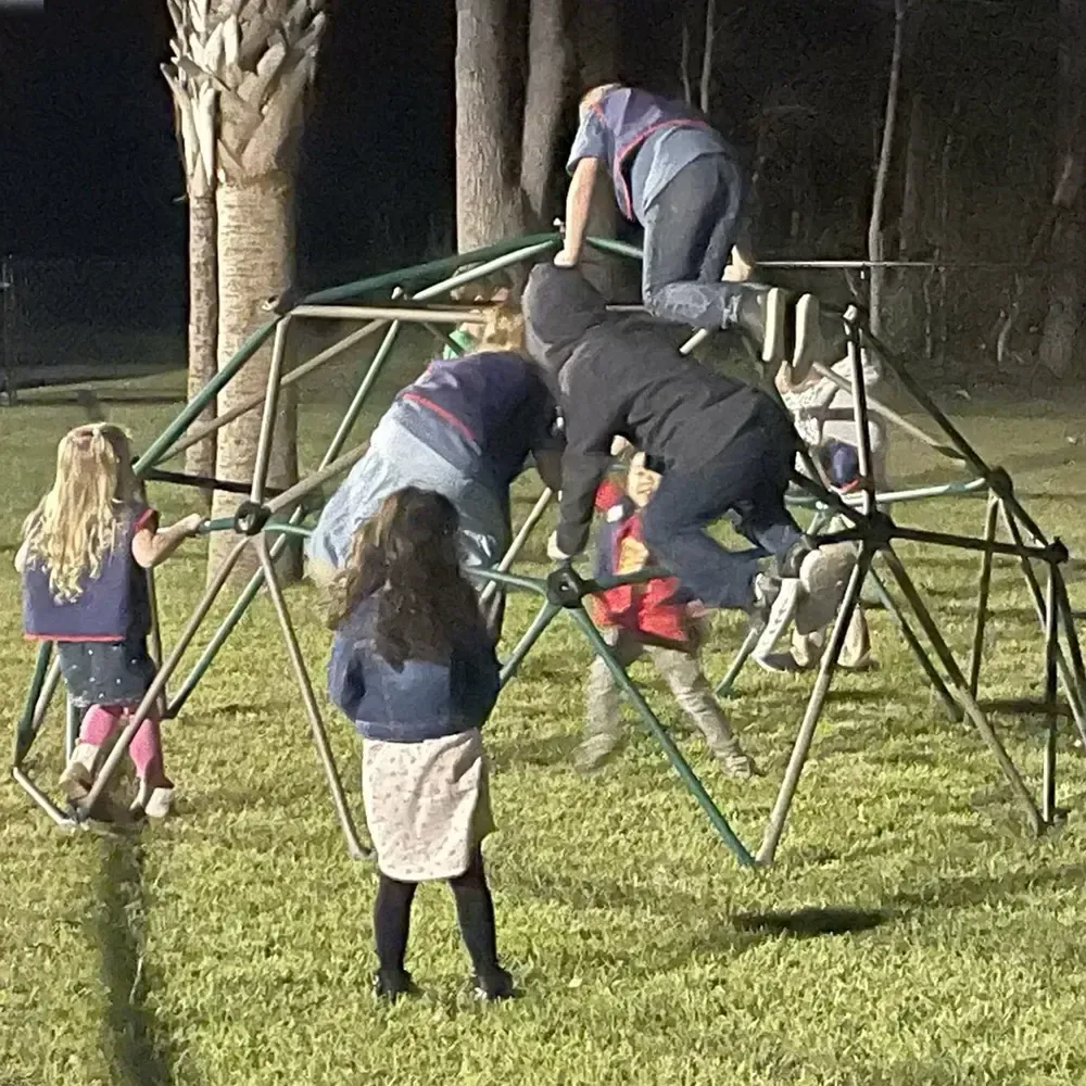 Children climbing a green dome playground at night. Others watch from the ground.