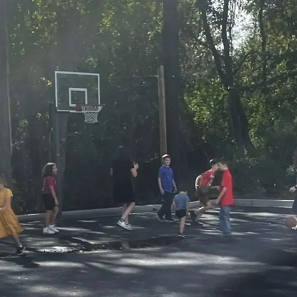 Children playing basketball outdoors on a paved surface with trees in the background.