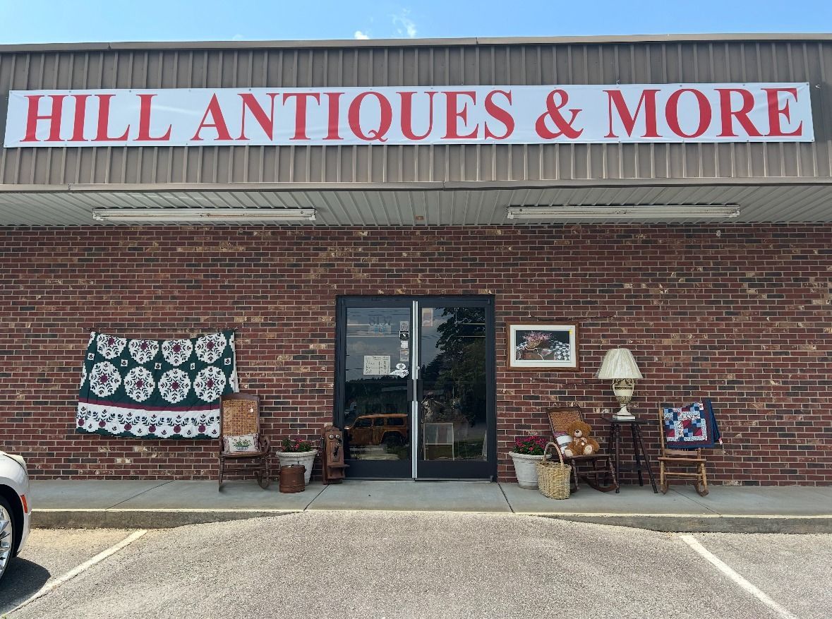 Brick storefront with “HILL ANTIQUES & MORE” sign, antiques displayed outside, and a glass entrance door.