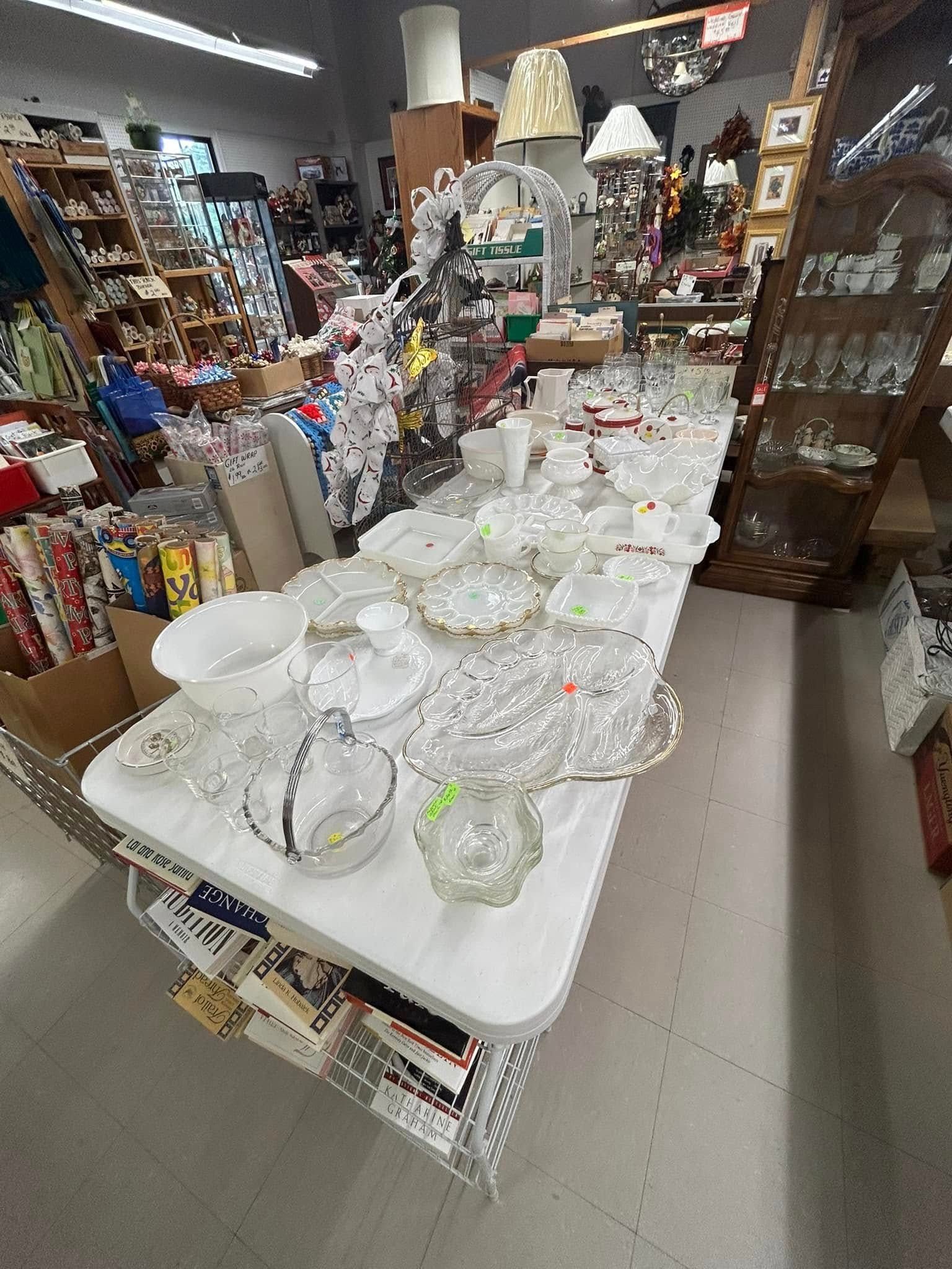 Glassware displayed on a table in a cluttered shop aisle, with shelves of assorted items in the background