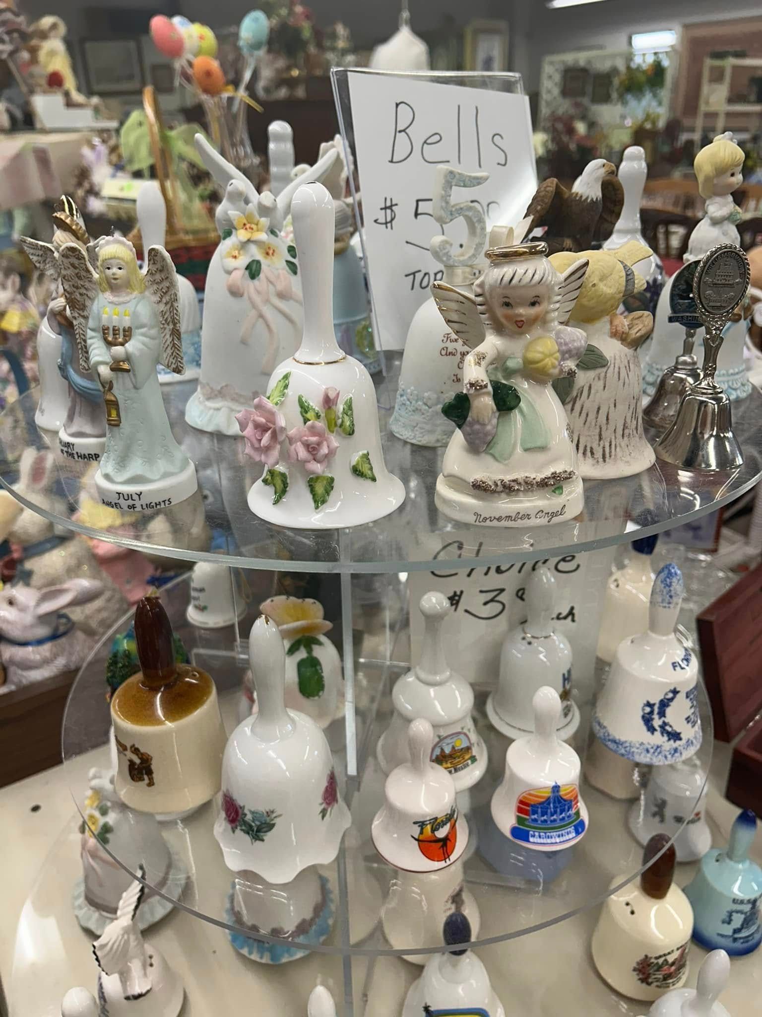 Shelf of decorative ceramic bells and figurines, many white with floral accents, in a shop display.