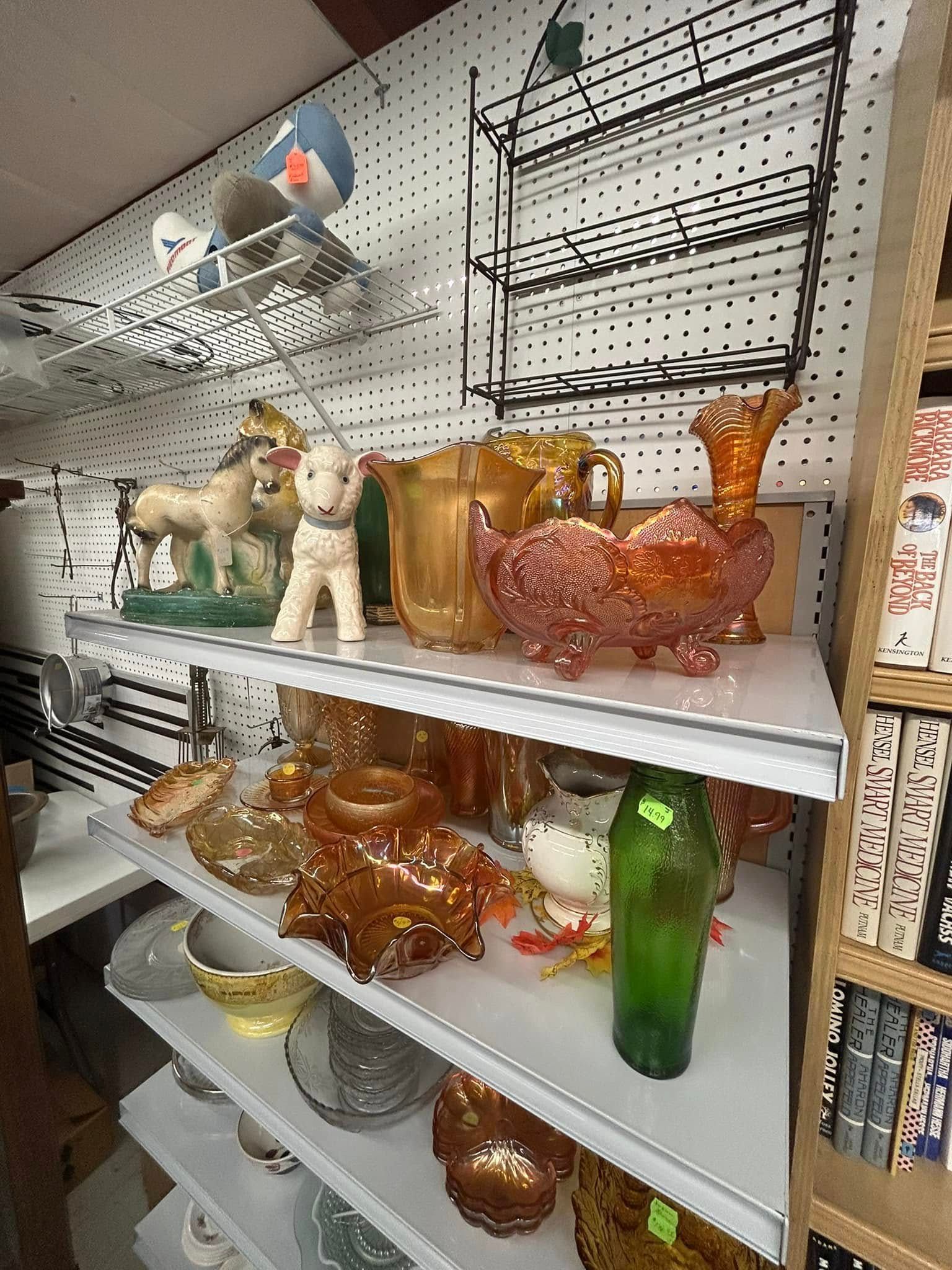 Table of glassware and dishes in a cluttered shop aisle, with shelves of assorted items around it