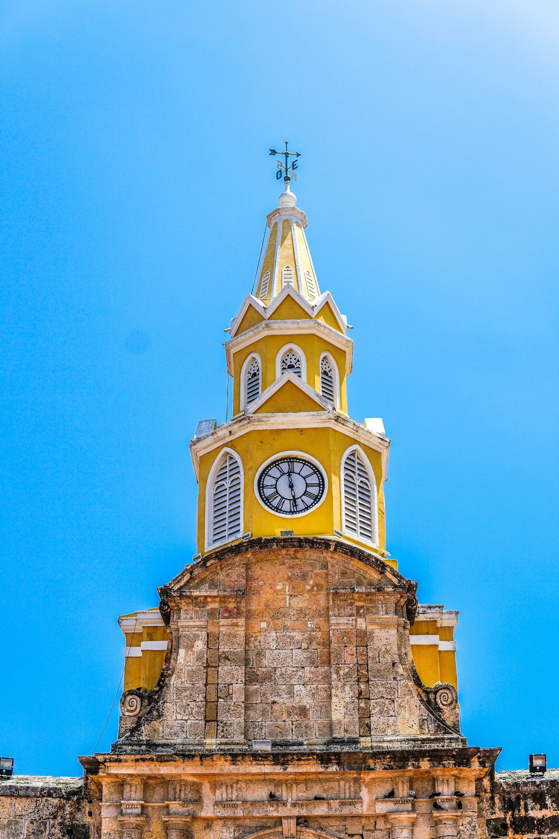 Una torre de reloj amarilla con una cruz en la parte superior contra un cielo azul.