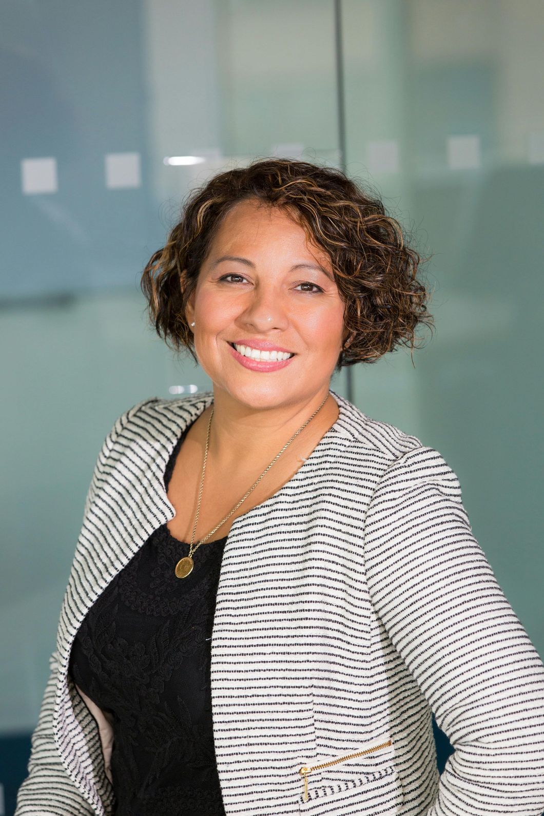 Woman in black dress and patterned jacket smiles, in front of a glass wall.