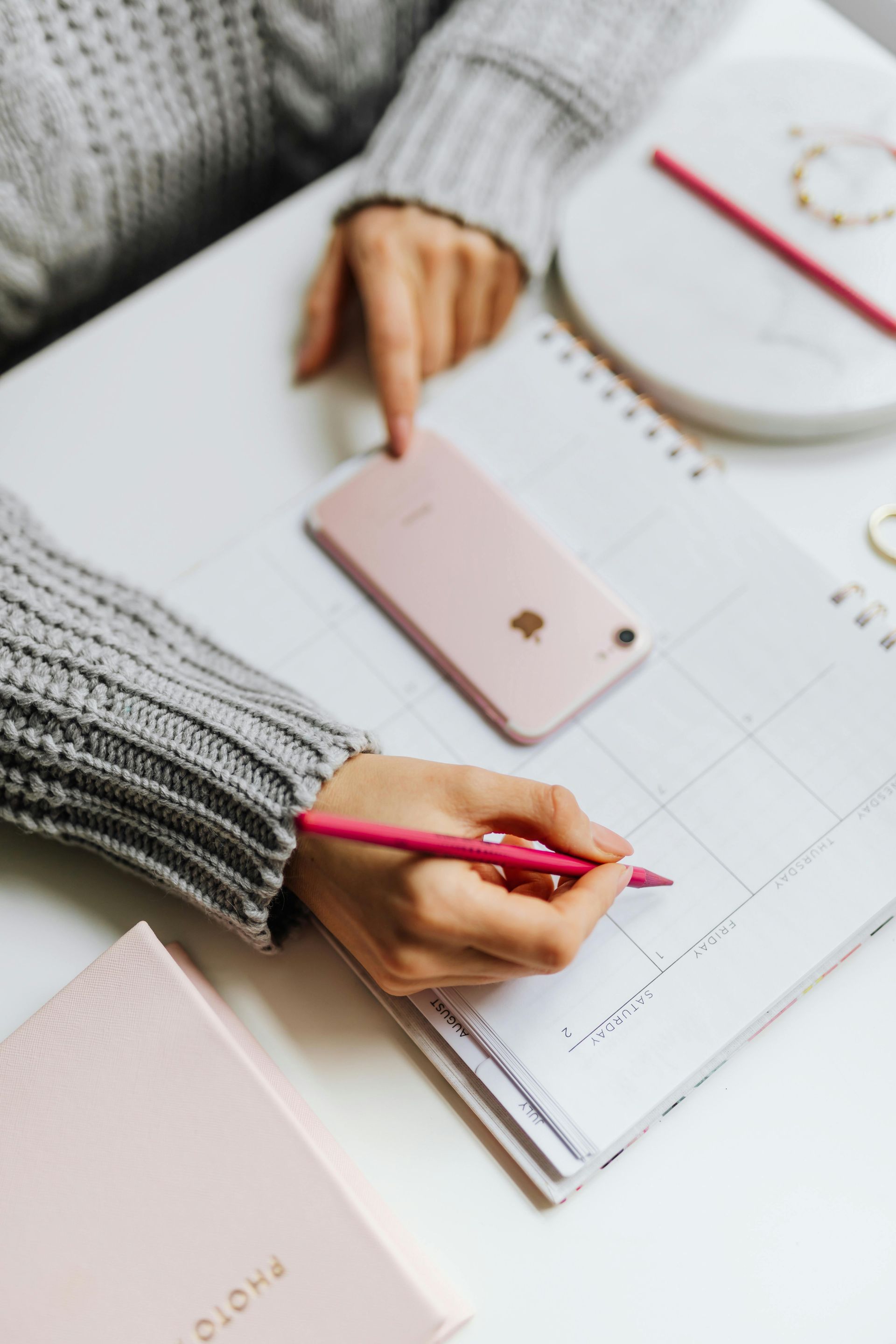Person writing in pink pen on calendar, with phone and accessories on a white table.