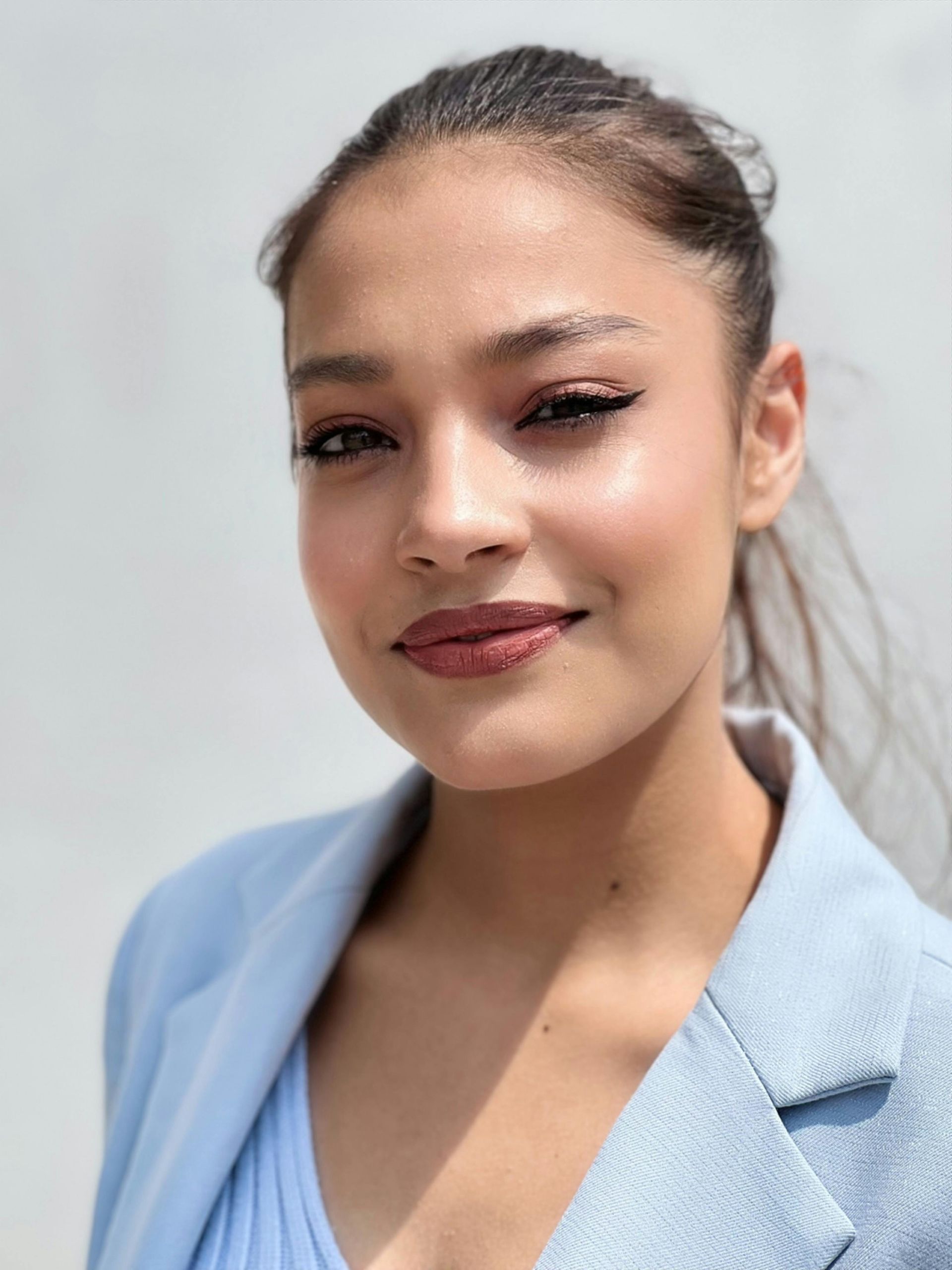 Woman with brown eyes and slicked-back hair smiles, wearing a blue blazer against a white background.