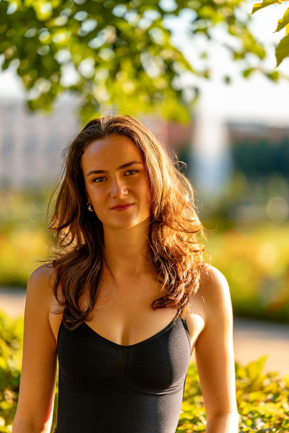 Woman with brown hair, wearing a black tank top, smiling outside in a sunlit park.