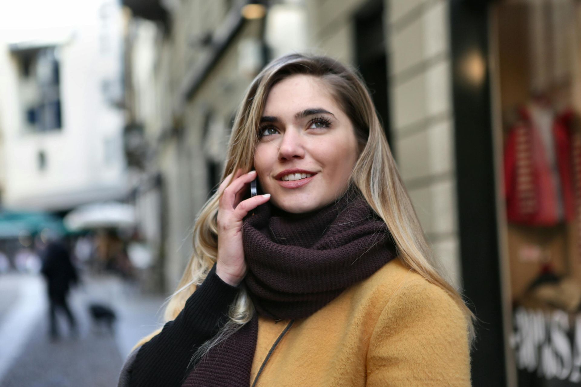 Woman on a phone call in an alley, wearing a brown scarf and yellow coat, smiling.